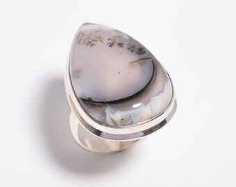 May include: A silver ring featuring a large, teardrop-shaped stone. The stone has a marbled appearance with shades of gray, white, and black, and a subtle, tree-like pattern. The ring's band is a simple, polished silver.