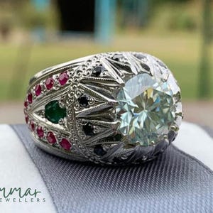 Peut inclure: Une bague en argent ornée d'une grosse pierre précieuse ronde et vert clair au centre. La bague est ornée de petites pierres précieuses rouges, vertes et noires. La monture présente des détails de filigrane complexes. Le logo Ammar Jewellers est visible.