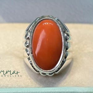 May include: A silver ring featuring an oval, vibrant orange gemstone. The ring has a detailed, textured setting around the stone and a wide band. The ring is displayed in a light blue jewelry box.