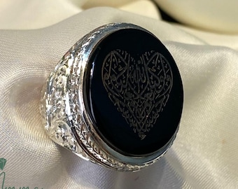 May include: A silver ring with an oval, black onyx stone. The stone features an engraved heart design with Arabic calligraphy. The ring has a detailed, floral pattern on the band. The Ammar Jewellers logo is in the bottom left corner.