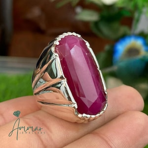 Op de afbeelding: Een zilveren ring met een grote, rechthoekige robijn edelsteen. De robijn heeft een dieprode kleur en is gefacetteerd om het licht te vangen. De band van de ring is van gepolijst zilver en de zetting heeft een decoratieve rand. De ring is een sieraad.