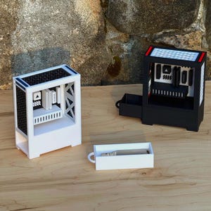 May include: Two miniature computer cases, one white and one black, sit on a wooden surface. The white case has a black top and blue accents, while the black case has a red accent. A small white rectangular box with a handle is in front of them.