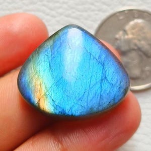 Puede incluir: Una piedra preciosa de labradorita pulida en forma de lágrima, con una iridiscencia azul y amarilla vibrante. La piedra se sostiene en una mano, con una moneda en el fondo, sirviendo como referencia de tamaño. La superficie de la piedra muestra patrones naturales.