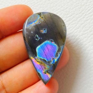May include: A teardrop-shaped labradorite gemstone with a polished surface. The stone displays a dark gray base color with iridescent flashes of blue, purple, and yellow. The gemstone is held in a hand against a white background.