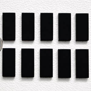 Può includere: Dieci oggetti rettangolari neri, simili a tessere del domino, sono disposti su una superficie bianca. Una moneta è posta accanto agli oggetti per la scala. Ogni oggetto ha una superficie liscia e riflettente. Per giochi.