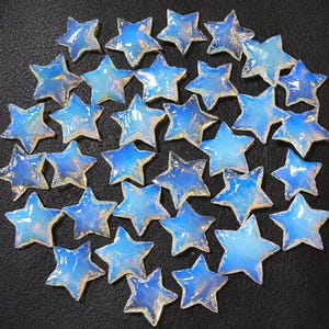 May include: A cluster of opalescent star-shaped beads. The beads display a translucent, iridescent sheen with hues of blue and white. The stars are closely arranged against a dark backdrop, creating a celestial appearance. Ideal for jewellery making and crafts.