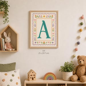May include: A framed print featuring the letter "A" in teal, surrounded by a floral border. The print is A4 size, measuring 21 x 29.7 cm. The artwork is displayed in a light-colored wooden frame, suitable for a nursery or children's room.