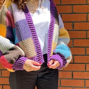 Könnte beinhalten: Ein farbenfroher Patchwork-Häkelcardigan in Lila-, Gelb-, Blau- und Beigetönen. Der offene Pullover hat einen dicken, gerippten lila Besatz und wird über einem weißen T-Shirt getragen. Die Ärmel sind lang und der Cardigan ist aus dickem Garn gefertigt.