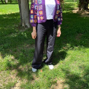 May include: A handmade, purple crochet cardigan with colorful square patterns. The cardigan has a V-neck and long sleeves. The person is wearing a white top, black pants, and white sneakers. The background is a green, grassy area with trees.
