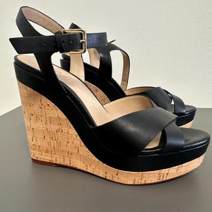 May include: Black wedge sandals with a cork platform and heel. The sandals have a leather upper, a crisscross strap design, and an ankle strap with a gold buckle. The platform is about 2.5 cm, and the heel is approximately 10 cm.