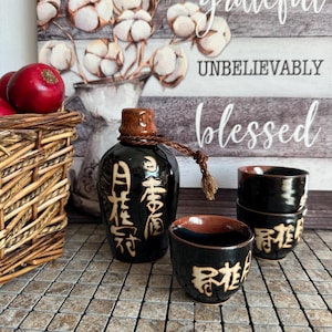 Könnte beinhalten: Ein Sake-Set aus schwarzer Keramik, bestehend aus einer Flasche mit braunem Hals und geflochtener Kordel sowie zwei passenden Bechern. Jedes Teil ist mit handgemalten beigen japanischen Schriftzeichen auf einer glänzend schwarzen Glasur versehen.