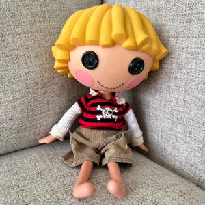Pode incluir: Uma boneca com cabelo moldado amarelo, olhos de botão pretos e bochechas circulares cor-de-rosa. Veste uma camisola às riscas vermelhas e pretas com um gráfico de caveira branca, mangas compridas brancas e calções de bombazina bege.