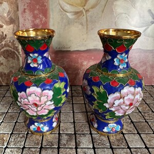 May include: A pair of cloisonné vases featuring a vibrant blue base with intricate floral patterns in pink, green, red, and light blue. Each vase has a gold-toned rim and base, with delicate metal wire detailing outlining the colourful enamel designs.