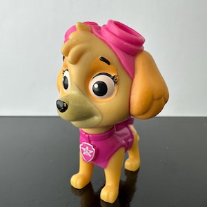 May include: A plastic Skye figurine from Paw Patrol. The figure is light tan with orange ears and a pink helmet and vest. It has large black eyes and a small black nose. The vest features a pink badge.