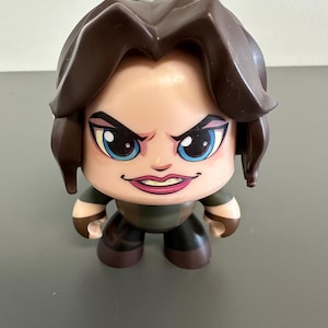 May include: A stylized figurine with a determined expression. The figure has brown hair, blue eyes, and is wearing a green and brown outfit. The figurine is standing upright.