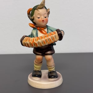 May include: A vintage figurine of a young person playing an accordion. The figurine is wearing a blue jacket, brown shorts, and a red scarf. The accordion is tan and orange. The figurine is standing on a round, off-white base.