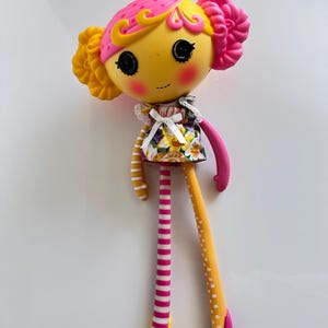 Puede incluir: Una muñeca colorida con cara amarilla, pelo rosa y vestido floral. La muñeca tiene ojos negros de botón, mejillas rosadas y una pequeña corona naranja. Una pierna es a rayas rosas y blancas, la otra amarilla con lunares blancos.