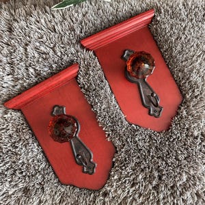 Könnte beinhalten: Zwei Wandhaken mit rechteckigen, roten Holzplatten und einer oberen Ablage. Jede Platte ist mit einem dunklen Metallbeschlag und einem facettierten, durchscheinenden bernsteinfarbenen Glasknauf ausgestattet, der als funktionaler Haken dient.