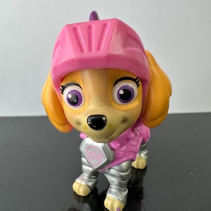 May include: A plastic Skye figurine from the Paw Patrol series. The figurine is in a standing position, wearing a pink helmet and armor. The dog has orange ears, big purple eyes, and silver-coloured legs.