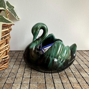 Peut inclure: Un cache-pot en céramique en forme de cygne avec une glaçure vert forêt profond et brillant, agrémenté de nuances vert noir. Le cygne présente un long cou courbé et des détails d'ailes moulés, avec une étiquette en papier bleu et blanc.