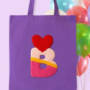 Puede incluir: Un bolso tote morado con una gran letra "B" texturizada y un diseño de corazón rojo. La letra está hecha de tela rosa, crema y amarilla. Un montón de globos de colores están en el fondo.