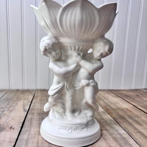 Peut inclure: Un vase en céramique blanc avec un motif de fleur de lotus au sommet, soutenu par deux chérubins. Le vase a une base ronde avec des détails décoratifs. Le design est élégant et classique, adapté à la décoration intérieure.