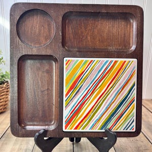 May include: A dark brown wooden serving tray with four compartments. One circular, two rectangular, and one square tile with a colourful diagonal stripe pattern. The tray is displayed on a black stand.