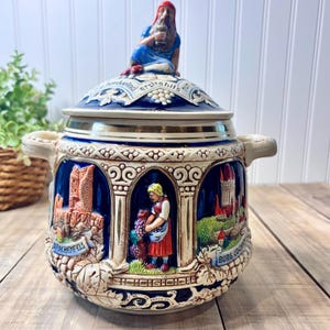 May include: A ceramic lidded stein with a blue and cream color scheme. The stein features relief scenes of castles and figures, with a seated figure on the lid. The stein has two handles and decorative trim.
