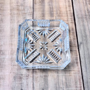 May include: Clear glass ashtray with a square shape and intricate starburst and geometric designs. The ashtray has a textured rim and a central floral pattern. The ashtray is sitting on a wooden surface.