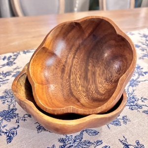 May include: Two wooden bowls with a unique, scalloped edge design. The bowls are made of a warm-toned wood, showcasing the natural grain. One bowl is nested inside the other, suggesting a serving or decorative set. The bowls sit on a blue and white floral patterned cloth.