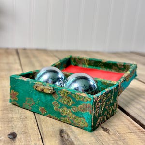 May include: Two polished chrome meditation balls rest inside a rectangular green fabric box. The box features a gold-toned metal latch and a vibrant red interior lining. The exterior fabric has a woven pattern with green and gold-colored threads.