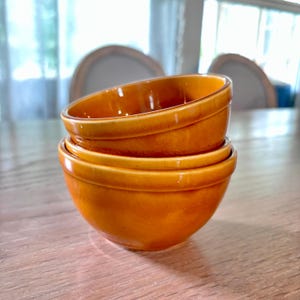 May include: A stack of three small, round, orange-glazed ceramic bowls. The bowls have a simple, classic design with a slightly flared rim and a smooth, glossy finish. They are stacked on top of each other on a wooden surface.