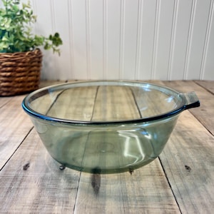 May include: A clear, olive-green glass mixing bowl with a black rim and a handle. The bowl is round and sits on three small feet. The bowl is empty and appears to be vintage.