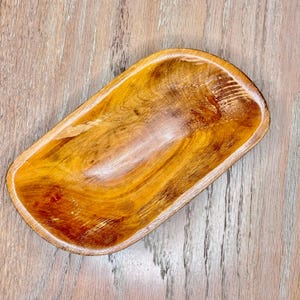 May include: A rectangular wooden bowl with rounded edges. The bowl is made of a light brown wood with a visible grain pattern. The interior of the bowl is smooth and polished, while the exterior has a slightly rougher texture.