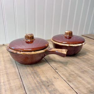 May include: Two brown ceramic soup bowls with lids. One bowl has a handle. The bowls have a dark brown glaze with a lighter brown rim and lid handle. The bowls are on a wooden surface.