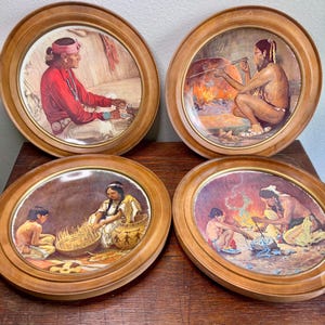 May include: Four round decorative plates with wooden frames. Each plate features a detailed painting of Native American figures engaged in various activities. The artwork includes vibrant colors and depicts scenes of daily life and cultural practices.