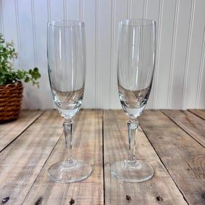 May include: Two clear champagne flutes with vertical etched lines. The flutes have a slender stem and a round base. The glasses are on a wooden surface with a white background.