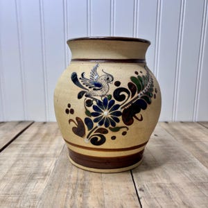 May include: A tan ceramic vase with a dark brown rim and base. The vase is decorated with a blue and white bird, blue flowers, and green and brown foliage. The vase has a round shape and a wide opening.