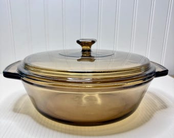 May include: A vintage amber-colored glass casserole dish with a matching lid. The dish has a round shape with two handles and a knob on the lid. The glass has a warm, transparent hue.