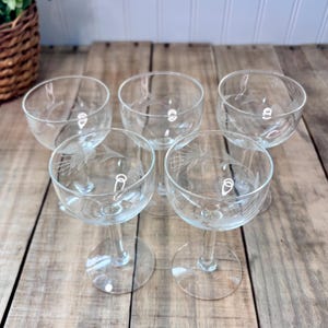 May include: Five clear champagne coupe glasses with etched floral designs. The glasses have a round bowl shape, a slender stem, and a circular base. The glasses are arranged on a wooden surface.