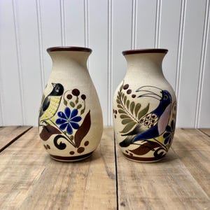 May include: Two ceramic vases with a beige speckled finish. Each vase features a hand-painted bird design with blue, yellow, and brown accents. The vases have a brown rim and are displayed on a wooden surface.
