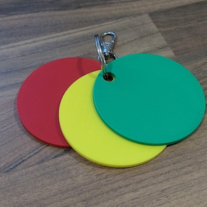 May include: Three circular tags in red, yellow, and green, connected by a silver metal clasp. The tags are layered, with the red tag at the bottom, the yellow in the middle, and the green on top. The tags are made of a smooth material.