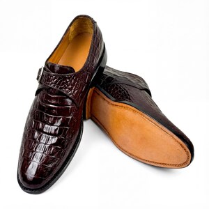 May include: A pair of brown leather dress shoes with a crocodile skin texture. The shoes have a buckle closure and a leather sole. The shoes are displayed on a white background.