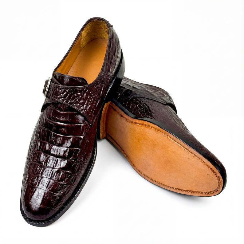 May include: A pair of brown leather dress shoes with a crocodile skin texture. The shoes have a buckle closure and a leather sole. The shoes are displayed on a white background.