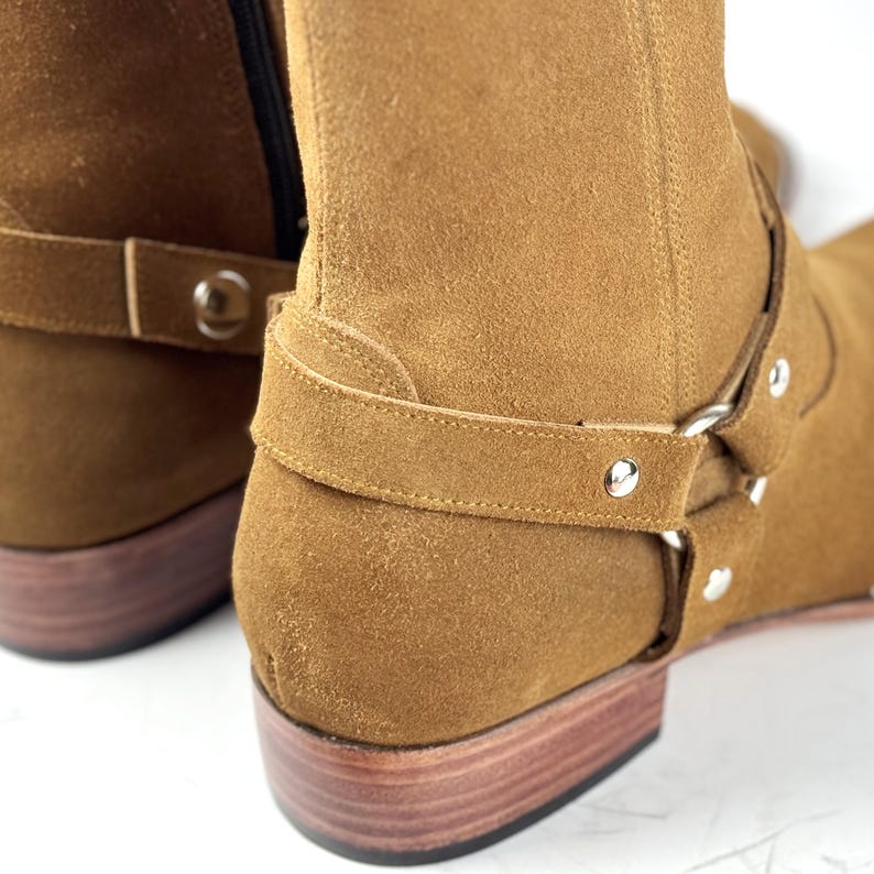 May include: Close-up of tan suede boots with a harness strap and silver-tone hardware. The boots have a stacked wooden heel and a side zipper. The suede has a soft, textured appearance.