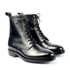 Puede incluir: Un par de botas de combate de cuero negro con diseño de cordones. Las botas tienen punta redondeada, tacón bajo y una lengüeta en la parte trasera. Las botas están sobre un fondo blanco.