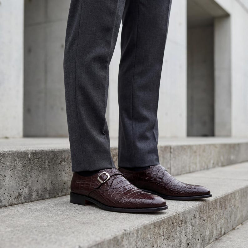 May include: A pair of brown leather dress shoes with a crocodile skin texture. The shoes have a buckle closure and are worn with gray dress pants. The shoes are on a concrete staircase.