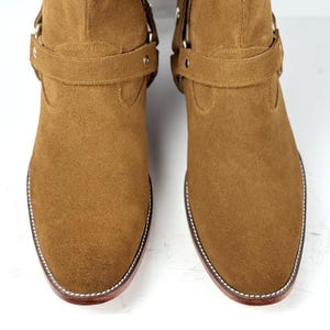 May include: A pair of brown suede boots with a pointed toe and a strap detail. The boots have a stitched sole and a low heel. The boots are made of a soft, textured material.