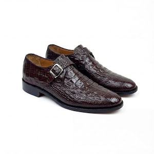 May include: A pair of brown leather dress shoes with a crocodile skin texture. Each shoe features a silver buckle closure and a rounded toe. The shoes are set against a white background.