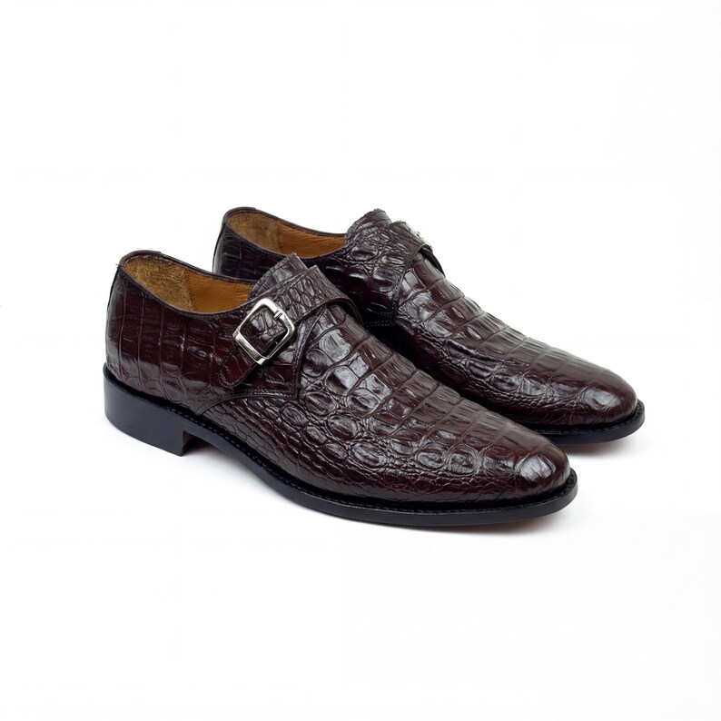 May include: A pair of brown leather dress shoes with a crocodile skin texture. Each shoe features a silver buckle closure and a rounded toe. The shoes are set against a white background.